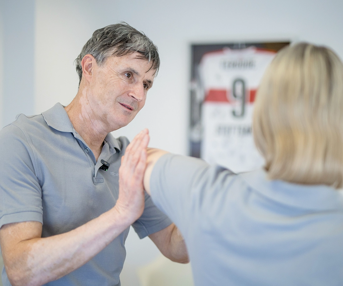 Sportheum_Sportklinik_Fellbach_Fachbereiche_Schulter_Ellenbogen_Dr_Mauch_tvb Dr. Mauch von der Sportheum Sportklinik Fellbach bei der Untersuchung der Schulter einer Patientin – im Hintergrund ein signiertes Trikot des VfB Stuttgart.