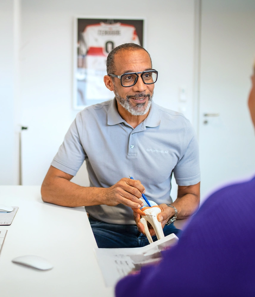 Sportheum_Sportklinik_Fellbach_Fachbereiche_vfb_Stuttgart_Diagnostik Arzt der Sportheum Sportklinik Fellbach erklärt einem Patienten mit einem anatomischen Knie-Modell eine Verletzung – im Hintergrund ein signiertes Trikot des VfB Stuttgart.